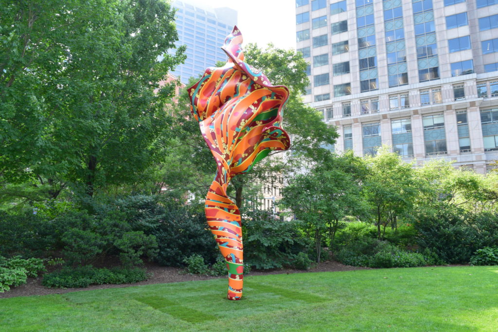 A large, orange, red and green sculpture that looks like a billowing piece of fabric sits on a bright green lawn. Framed by trees, the background reveals large urban buildings surrounding the artwork.