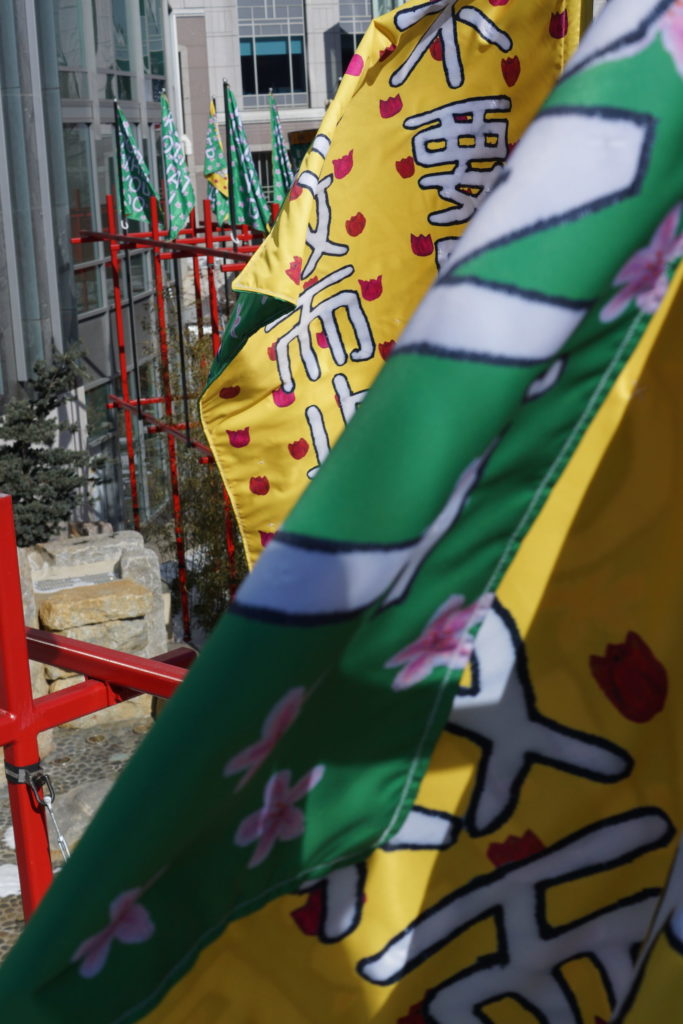 In the foreground, two banners wave vigorously, revealing the texture and details of banners, such as the red and pink flower details on the yellow and green backgrounds, respectively. In the background, more green banners sit restfully atop a red structure.