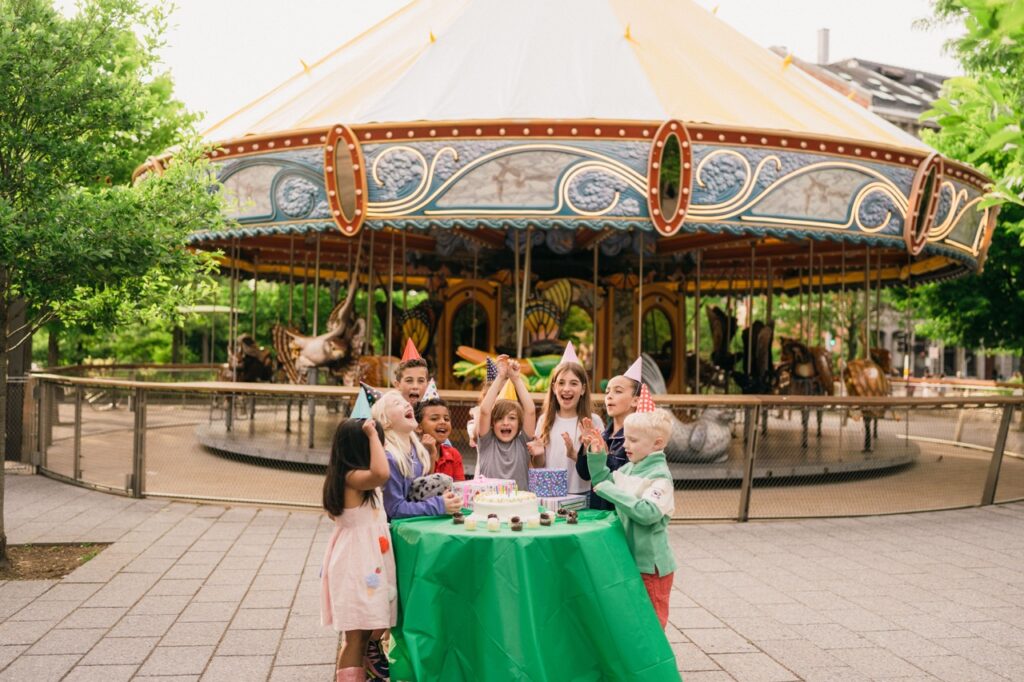 A group of childre are gathered around a round table with a green tablecloth. They are wearing party hats and there is a white cake on the table. In the background is a colorful carousel.