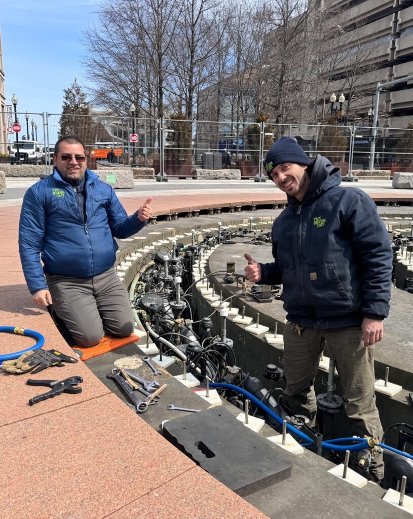 Two men perform maintenance on an in-ground fountain. Wires and tools can be seen on the ground next to them. They are smiling at the camera.