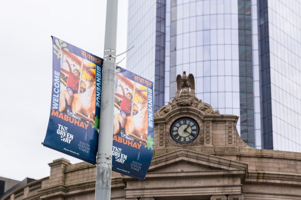 Two blue banners on a lamp post show an image of a boy riding a fox on the Greenway Carousel. Around the image is the word "Welcome" in 4 different languages. Below the image are two logos. Behind the banner is a clock at the entrance to South Station.
