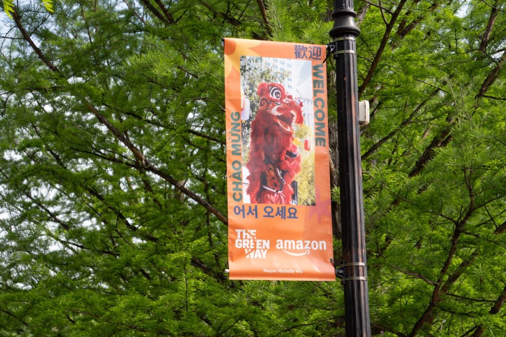 An orange banner on a lamp post shows an image of a traditional Chinatown lion dance with a red lion costume. Around the image is the word "Welcome" in 4 different languages. Below the image are two logos.