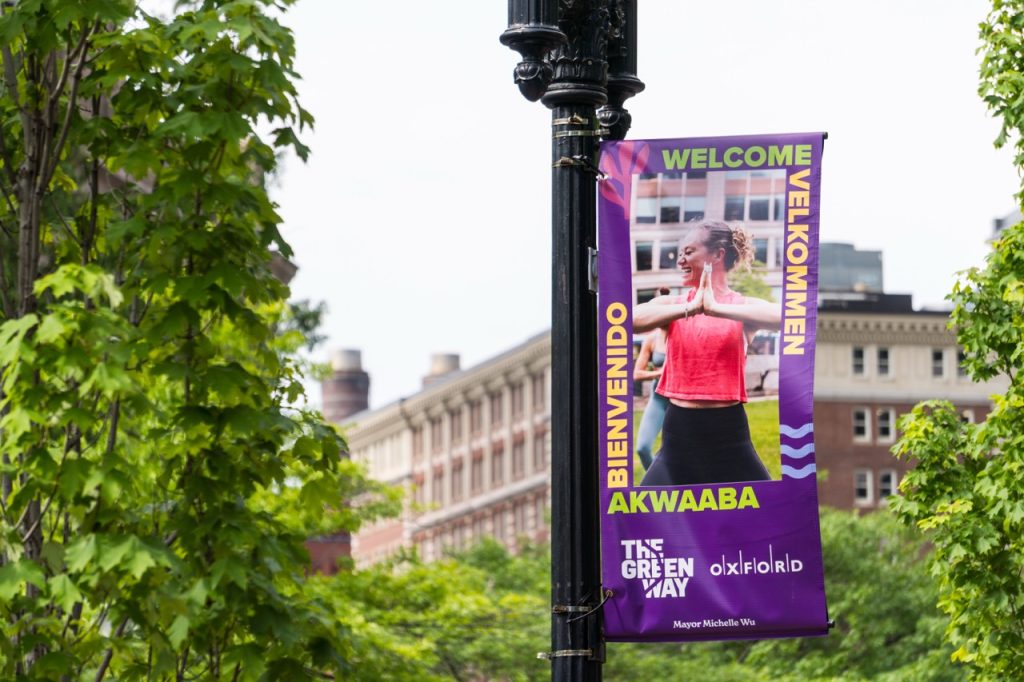 A purple banner on a lamp post shows a photo of a woman in a pink top doing a fitness post. Around the image is the word "Welcome" in 4 different languages. Below the image are two logos.
