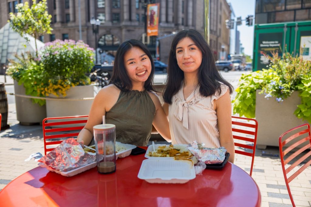 Two young women are sitting at a red table in an outdoor city setting. There are takeout boxes of food on the table in front of them.