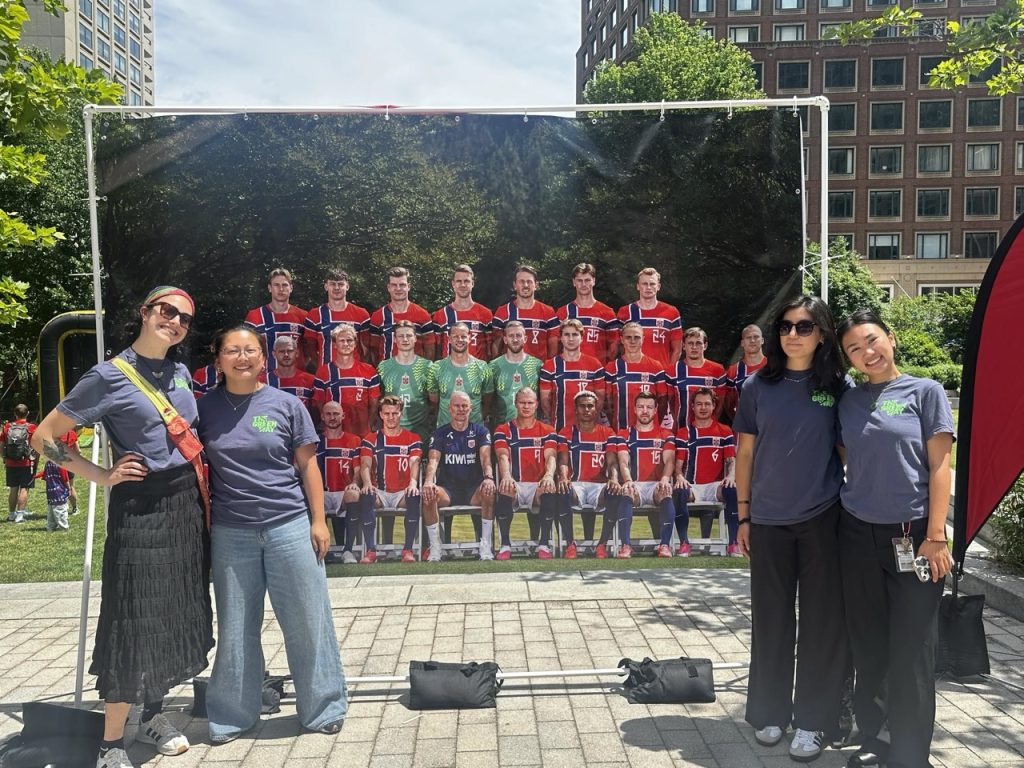 4 young adults stand in front of a large photo of the Norwegian soccer team.