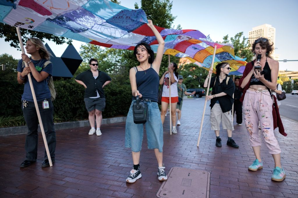 A group of people stands holding a large quilt-like sheet over their heads. They are on a sidewalk in a city setting.