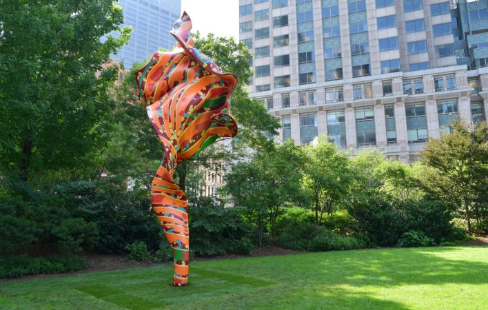 A large, orange, red and green sculpture that looks like a billowing piece of fabric sits on a bright green lawn. Framed by trees, the background reveals large urban buildings surrounding the artwork.