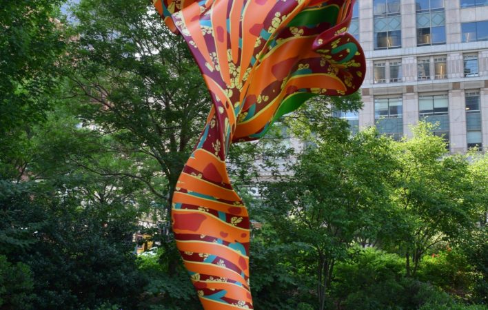 A large, orange, red and green sculpture that looks like a billowing piece of fabric sits on a bright green lawn. Framed by trees, the background reveals large urban buildings surrounding the artwork.