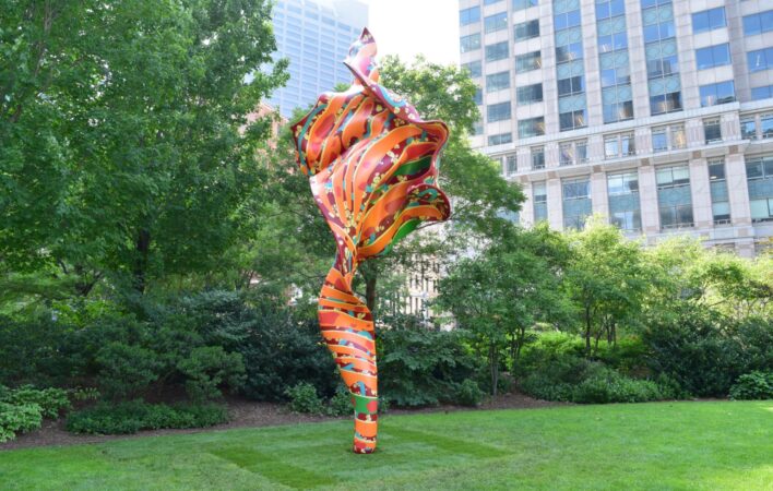 A large, orange, red and green sculpture that looks like a billowing piece of fabric sits on a bright green lawn. Framed by trees, the background reveals large urban buildings surrounding the artwork.