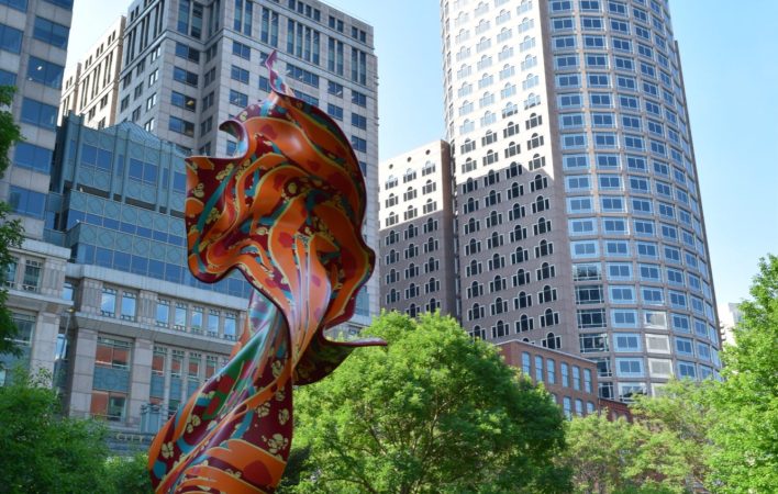 A large, orange, red and green sculpture that looks like a billowing piece of fabric sits on a bright green lawn. Framed by trees, the background reveals large urban buildings surrounding the artwork.