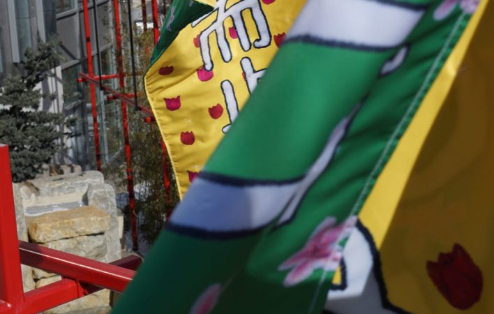 In the foreground, two banners wave vigorously, revealing the texture and details of banners, such as the red and pink flower details on the yellow and green backgrounds, respectively. In the background, more green banners sit restfully atop a red structure.