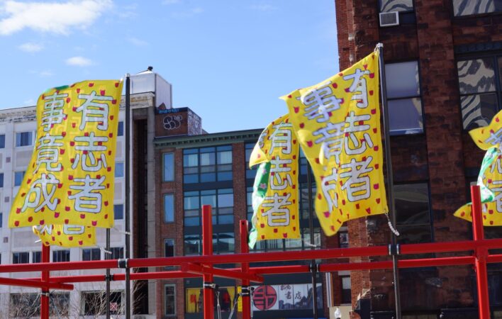 Four rectangular banners billow in the wind at the top of a red steel structure. Large white Chinese characters on the banners reads "IF THERE'S A WILL THERE'S A WAY" on a yellow background. The background features brick facades and a blue sky with one cloud.