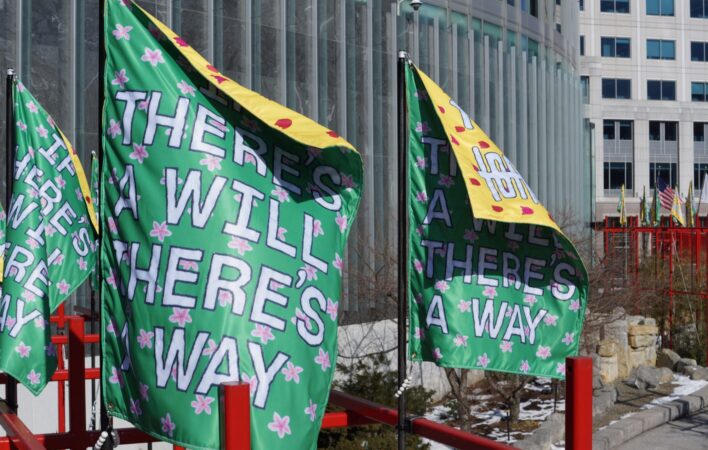 Three rectangular banners billow in the wind at the top of a red steel structure. Large white text on the banners reads "IF THERE'S A WILL THERE'S A WAY" on a green background. The corners of the banners reveal Chinese characters on a yellow background on the opposite side of the banners.