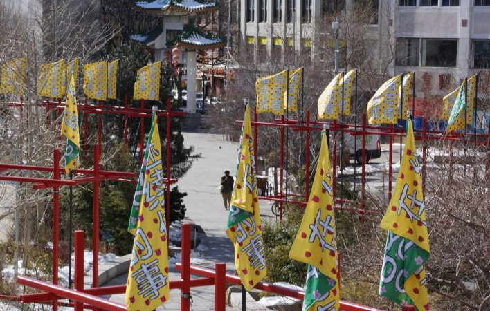 Four yellow and green banners drape restfully around their poles atop a red structure. In the background, numerous yellow banners billow in the wind, framing a sidewalk and the Chinatown gate and one distant figure walking down the path towards the gate.