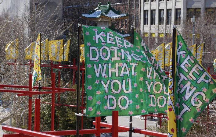 A rectangular banner is billowing horizontally to the pole supporting it, clearly displaying the text "KEEP DOING WHAT YOU DO" on a green background dotted with pink flowers. An identical banner to the right is folded in half in the wind. In the background, countless yellow banners billow in the wind and the Chinatown Gate fill the frame.